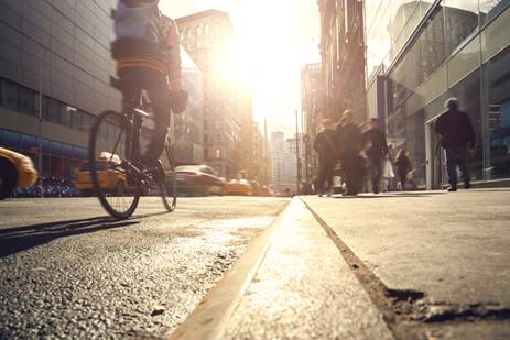A cyclist rides along a city street, alongside pedestrians and yellow taxis. The bright sunlight washes out the buildings in the background.